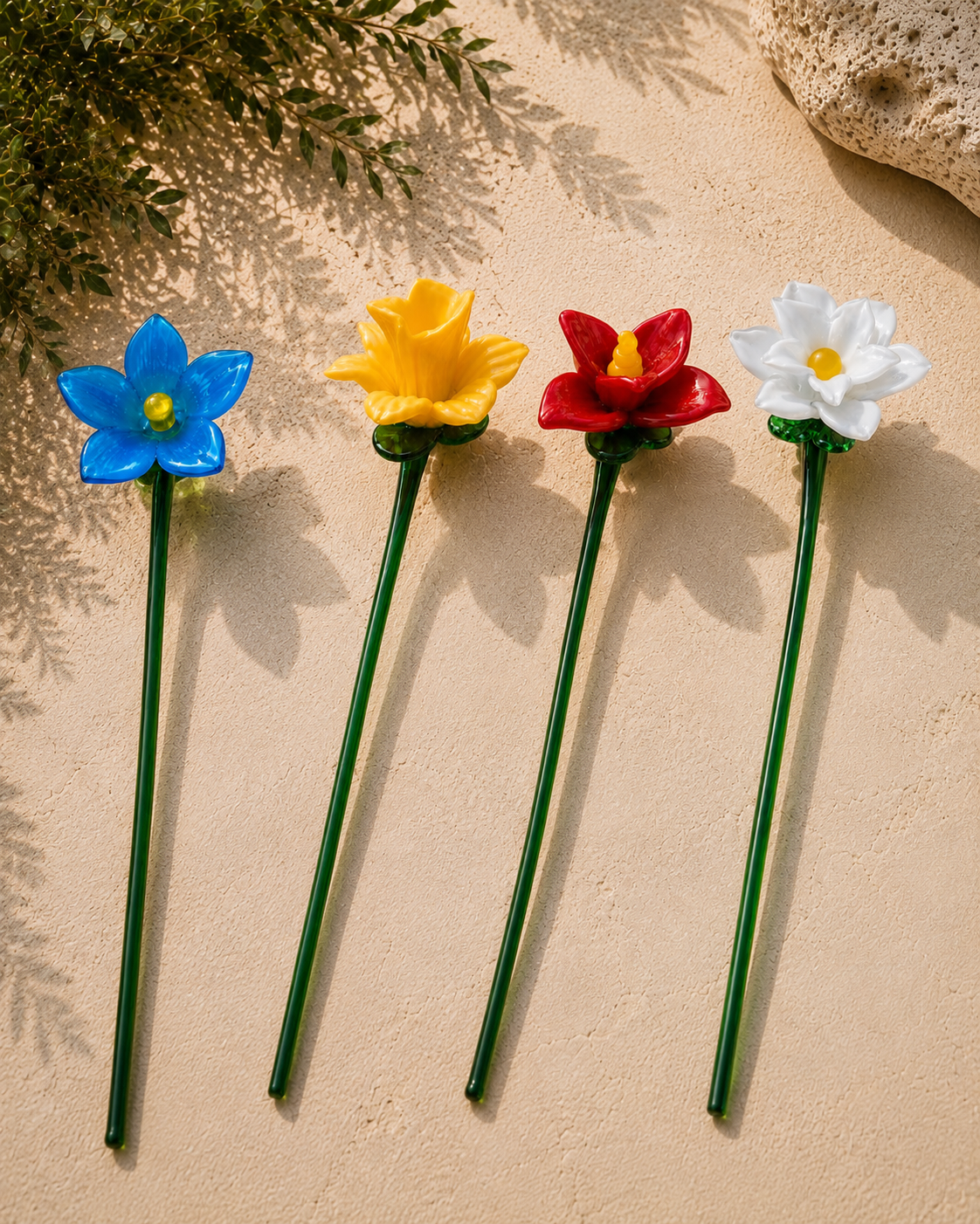 Four glass flowers side by side on limestone: blue, yellow, red and white.