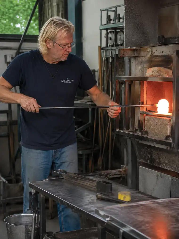 A glassblower in the workshop heats a glowing glass bulb at the end of a blowpipe in the furnace.