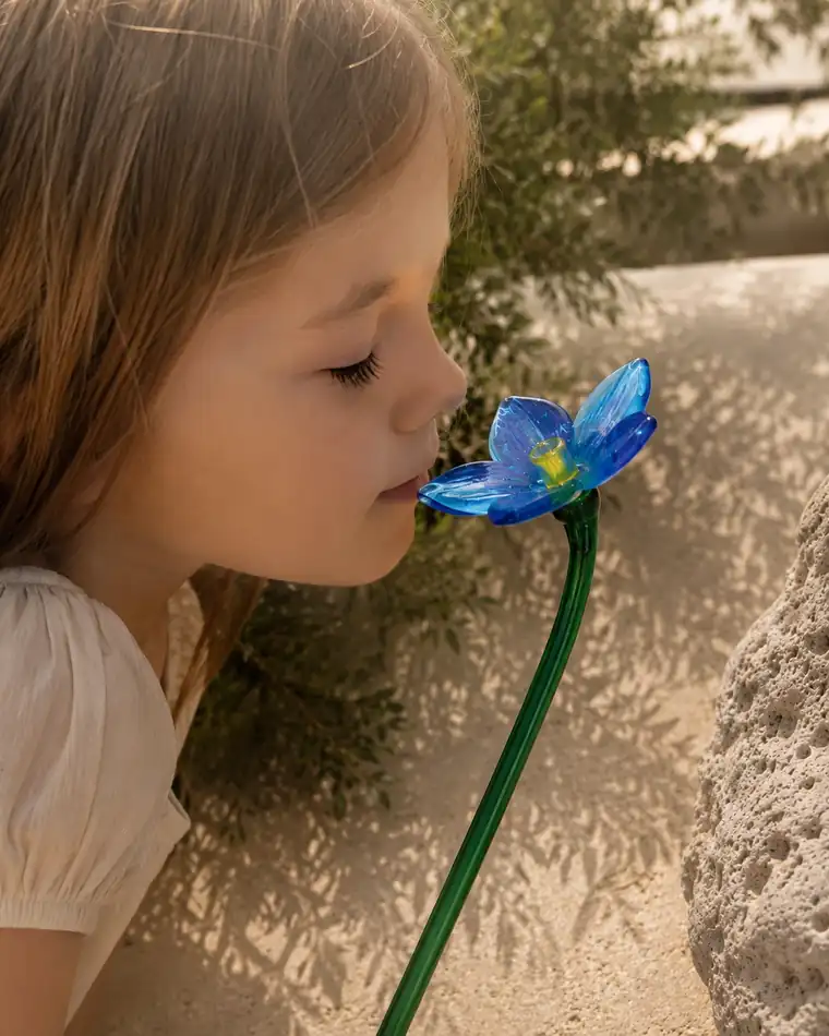 A little girl bends down to a pale blue glass flower and smells it.